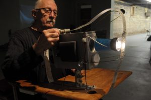 The image is a colour photograph of David Bobier, with his hair pulled back in a ponytail, wearing a black sweater and glasses. His right hand is extended and adjusting a Vibro-projector. 
