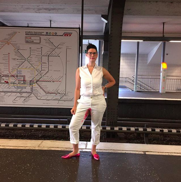 A photo of Vanessa wearing all white, with one hand on her hip. She has hot pink Menstrual Accessory between her legs, and hot pink shoes. She's standing on a metro platform, with the tracks behind her.