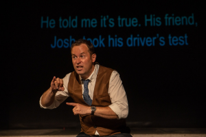 Chris Dodd performing Deafy at the SummerWorks Performance Festival. Chris wears a white shirt, tan vest and blue tie and is signing animatedly to the audience. Surtitles displayed behind him read "He told me it's true. His friend, Josh, took the driver's test."