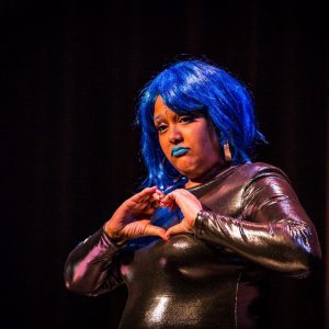 A photo of Gaitrie wearing a blue wig and blue lipstick. She is wearing a metallic bodysuit and signing.